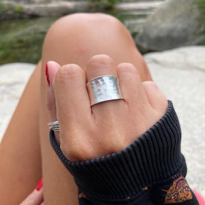 Peut inclure: Une large bague en argent martelé est portée au doigt. La bague a une surface texturée avec une forme légèrement irrégulière. La personne porte un pull noir avec un motif coloré.