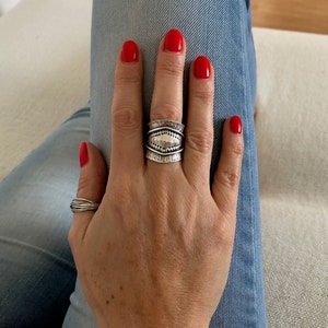 May include: A hand with red nail polish displays two silver rings. The larger ring has a textured design with a central oval shape. The smaller ring is a simple band of intertwined silver wires. The hand rests on blue denim.