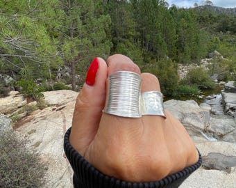 Könnte beinhalten: Zwei silberne Ringe an einer Hand mit rotem Nagellack. Die Ringe haben einzigartige Designs: einer mit gerippter Textur und der andere mit einem geätzten Muster. Der Hintergrund zeigt eine Naturlandschaft mit Bäumen und einem Bach.