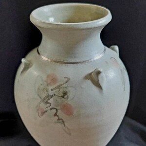 May include: A pale green ceramic vase with a rounded body and a narrow neck. The vase features two small handles and a floral design in pink and brown. A silver band encircles the neck.