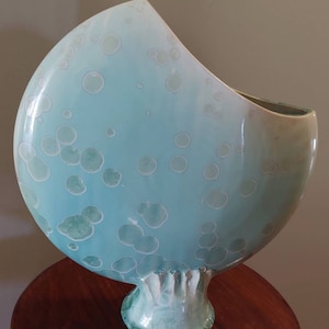 May include: A large, ceramic vase with an unusual, asymmetrical form. The vase is mainly light blue with a speckled pattern of lighter blue and white circles. The base is a textured, light green colour, and it rests on a dark brown wooden surface.
