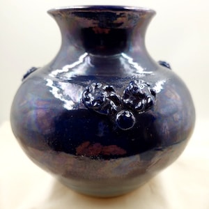 May include: A dark blue ceramic vase with a pearlescent finish. The vase has a rounded body, a narrow neck, and decorative raised floral-like embellishments. The vase is approximately 20 cm tall.
