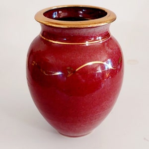 May include: A small, red ceramic vase with a gold rim and gold accents. The vase has a rounded body and a slightly flared opening.