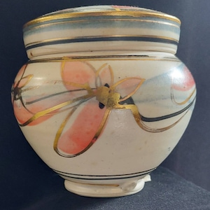 May include: A small, round, lidded ceramic jar with a floral design. The jar is primarily white with pink, gold, and black accents. The lid has a gold rim and blue and black stripes. The jar has a small foot.