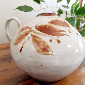 May include: A large, off-white ceramic jug with a rounded body and a handle. The jug is decorated with brown leaf-like designs. The rim and the handle are also brown. The jug is sitting on a wooden surface.