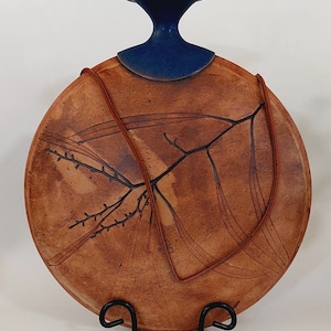 May include: A round, brown ceramic vase with a blue top and a brown leather strap. The vase features a design of a tree branch with leaves. The vase is on a black stand.