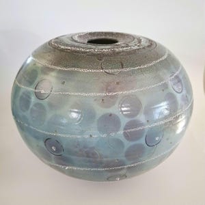 May include: A round ceramic vase with a light blue glaze and a pattern of white circles and dots. The vase has a textured surface and a slightly iridescent finish.