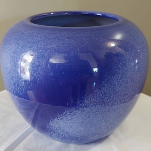May include: A round, blue ceramic vase with a speckled texture. The vase has a wide opening and a glossy finish. The colour transitions from a deep blue to a lighter, speckled blue near the base. A decorative item.