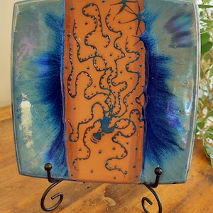May include: A square ceramic plate with a blue and orange glaze. The plate features a black design of a stylised octopus on the orange side. The plate is on a black metal stand.