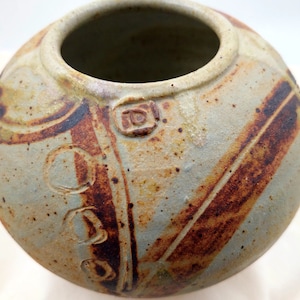May include: A handmade ceramic vase, round in shape with a wide opening. The vase has a light blue-grey base with brown and rust-coloured decorative stripes and circular patterns. A small, square, embossed design is visible.