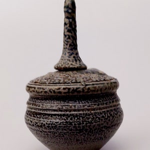 May include: A small, handcrafted ceramic jar with a lid. The jar is dark grey with a textured surface and horizontal etched lines. The lid has a tall, textured handle. The overall shape is rounded and bulbous.