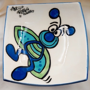 May include: Square ceramic dish featuring a cartoon butterfly design. The butterfly is rendered in blue and green with black outlines, on a white surface. The dish has a blue border and includes handwritten text in the upper left corner.