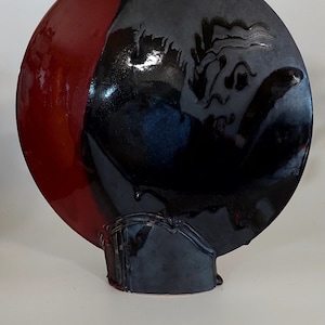 May include: A ceramic vase with a unique design. The vase is a combination of red and black glossy glaze. The vase has a circular shape with a cut-out at the top and a rectangular base. The black section has a textured design.