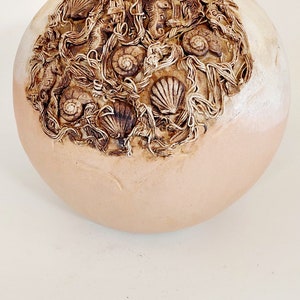 May include: A round, beige ceramic vase with a textured surface. The vase is decorated with a brown design of seashells, seaweed, and seahorses.