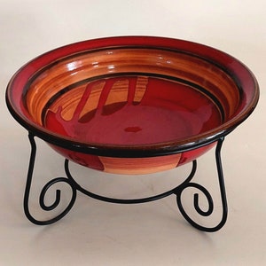 May include: A red and brown ceramic bowl with a decorative design sits on a black metal stand with three legs.