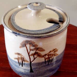 May include: A white ceramic jar with a lid, featuring a blue and brown tree design. The jar has a blue and white rim and a small spout on the lid.