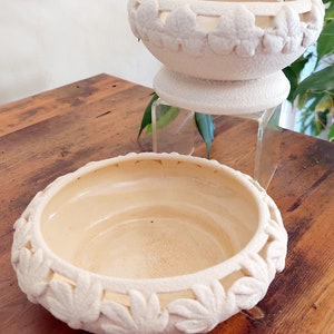 May include: Two cream ceramic bowls. The top bowl is on a pedestal. Both bowls have a textured surface and a leaf pattern around the rim. The bowls are on a wooden surface. The bowls are likely for decorative purposes.
