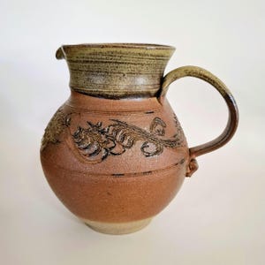May include: A brown ceramic jug with a green glaze and a carved design of leaves. The jug has a wide mouth and a handle.