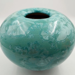 May include: A teal ceramic vase with a white glaze that creates a snowflake-like pattern. The vase has a round shape and a wide opening.