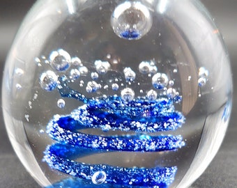May include: A clear glass paperweight with a blue spiral design and embedded air bubbles. The spiral is a vibrant blue with white speckles, creating a striking contrast against the clear glass. The paperweight is spherical.