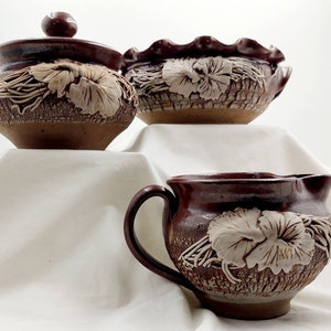 May include: A set of three brown pottery pieces with white floral designs. The set includes a creamer, a sugar bowl with a lid, and a bowl.