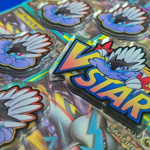 May include: A set of five acrylic charms featuring a Pokemon character with a VSTAR design. The charms are clear and have a glossy finish. The character is depicted in a blue, purple, and white color scheme. The text "VSTAR" is printed in a rainbow gradient.