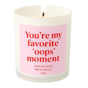 May include: A white candle in a frosted glass jar with a lit wick. The candle has a pink label with the text "You're my favorite 'oops' moment". The candle is made with 100% soy wax and weighs 300g.