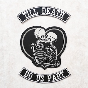 Könnte beinhalten: Zwei Skelette in einem herzförmigen Rahmen, die sich küssen. Der Text "Till Death Do Us Part" ist auf zwei schwarzen Bannern über und unter dem Herzen geschrieben.