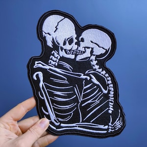 Könnte beinhalten: Gestickter Aufnäher mit zwei weißen Skeletten, die sich umarmen und küssen, vor einem schwarzen Hintergrund.