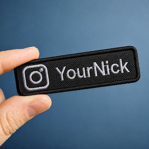 May include: A rectangular black embroidered patch featuring a white Instagram camera icon on the left side and the white text "YourNick" on the right side, with a stitched border surrounding the entire design on a dark fabric base.