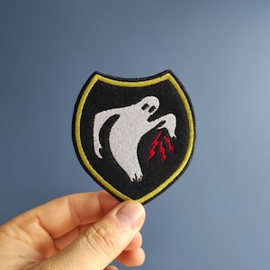 May include: An embroidered patch featuring a white ghost figure holding red lightning bolts. The patch is black with a yellow border and shaped like a shield.