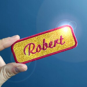 Peut inclure: Une étiquette de nom en paillettes dorées avec une bordure brodée rouge. Le nom "Robert" est écrit en lettres cursives rouges.