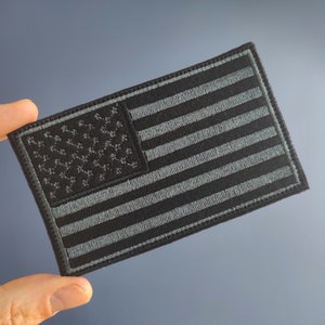 Op de afbeelding: Een geborduurde Amerikaanse vlagpatch in zwart en grijs. De vlag is gestikt met wit garen op een zwarte stoffen achtergrond.