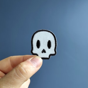 Op de afbeelding: Een witte schedelpatch met zwarte contouren, met twee oogkassen en een neus. De patch wordt in een hand tegen een blauwe achtergrond gehouden.