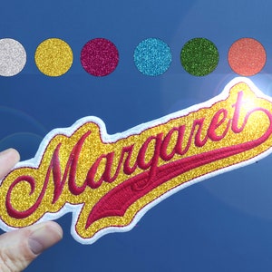 Peut inclure: Un patch thermocollant en paillettes dorées avec le nom "Margaret" en lettres pailletées rouges. Le patch est en écriture cursive et a une bordure blanche.