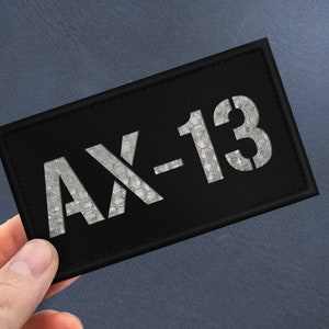 Könnte beinhalten: Schwarzer Stoff-Patch mit weißem Text "AX-13".