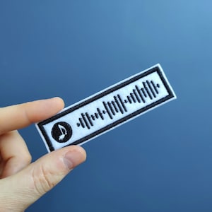 May include: White fabric patch with a black musical note and a black and white barcode-like pattern. The patch is held by a hand against a blue background.