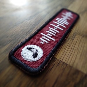 K&ouml;nnte beinhalten: Ein roter Stoff-Patch mit einem wei&szlig;en Soundwellen-Design und einem wei&szlig;en Kreis mit einem schwarzen Symbol in der Mitte. Der Patch hat schwarze N&auml;hte an den R&auml;ndern.