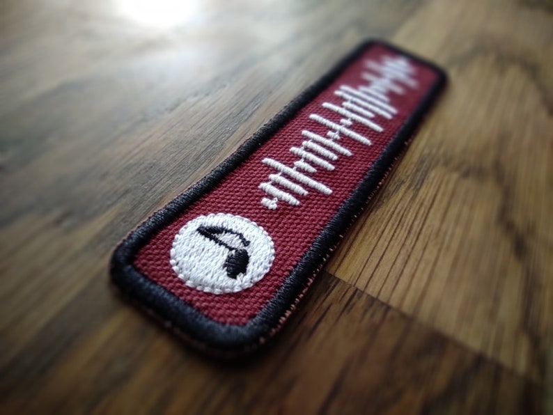 K&ouml;nnte beinhalten: Ein roter Stoff-Patch mit einem wei&szlig;en Soundwellen-Design und einem wei&szlig;en Kreis mit einem schwarzen Symbol in der Mitte. Der Patch hat schwarze N&auml;hte an den R&auml;ndern.