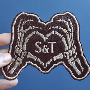 May include: A brown leather patch with two white skeleton hands forming a heart shape. The heart has the initials "S&T" embroidered in white.