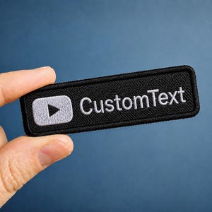 May include: A rectangular black embroidered patch featuring a white play button icon on the left side and the white text CustomText on the right side. The patch has a stitched border and a textured fabric surface, designed for attachment to clothing.