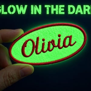 Pode incluir: Um emblema bordado de formato oval com um fundo verde neon que brilha no escuro, uma borda vermelha costurada e o nome Olivia bordado em letras cursivas vermelhas. O texto GLOW IN THE DARK aparece em verde acima do emblema.