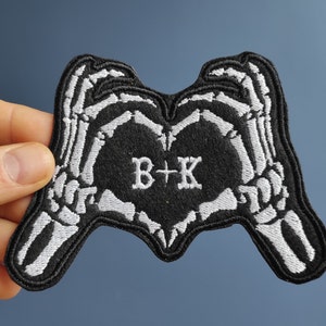 Könnte beinhalten: Schwarzer gestickter Patch mit zwei Skeletthänden, die eine Herzform bilden. Das Herz ist mit schwarzem Stoff gefüllt und trägt den Text "B+K" in weißer Stickerei.