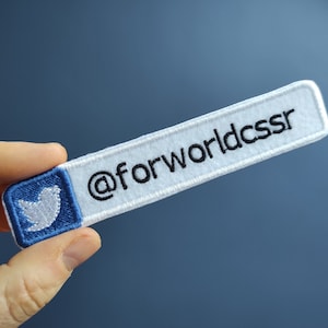 Könnte beinhalten: Weißer gestickter Aufnäher mit einem blauen Vogel-Symbol und dem Text "@forworldcssr" in Schwarz.