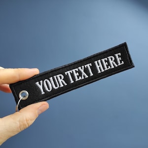 May include: Black fabric keychain with a silver metal ring and the text "YOUR TEXT HERE" embroidered in white.