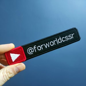 Könnte beinhalten: Schwarzer und roter gestickter Aufnäher mit einem weißen Play-Button und dem Text "@forworldcssr".