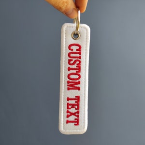 May include: White fabric keychain with a red embroidered text that says "CUSTOM TEXT".
