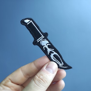 Può includere: Toppa ricamata nera a forma di coltello con contorno bianco. Il coltello è un design stilizzato con una lama e un manico.