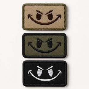Könnte beinhalten: Drei rechteckige bestickte Aufnäher mit einem schelmischen Smiley-Gesicht. Der obere Aufnäher ist beige mit dunkelbraunen Nähten, der mittlere ist olivgrün mit schwarzen Nähten und der untere ist schwarz mit weißen Nähten.