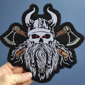 Könnte beinhalten: Gestickter Patch eines Wikingerkriegers mit einem Schädelgesicht, langem weißem Bart und zwei gekreuzten Äxten. Der Patch ist schwarz mit weißen und orangefarbenen Details.